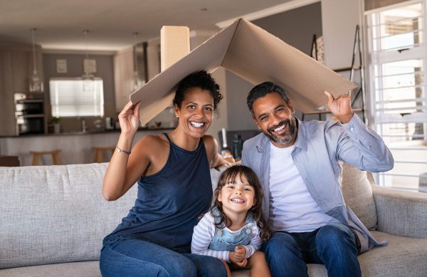 Portrait of smiling family sitting on couch holding cardboard roof and looking at camera. African and indian parents with daughter holding cardboard roof over heads while sitting on sofa in new home